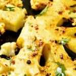 Grilled cauliflower steaks seasoned with spices and herbs, served on a black plate with garlic, parsley, and red pepper flakes on a dark rustic surface.