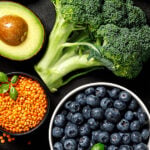 Colorful assortment of heart-healthy foods including fresh fruits, vegetables, legumes, seeds, and leafy greens such as broccoli, blueberries, strawberries, avocado, spinach, carrots, and tomatoes arranged on a dark background.